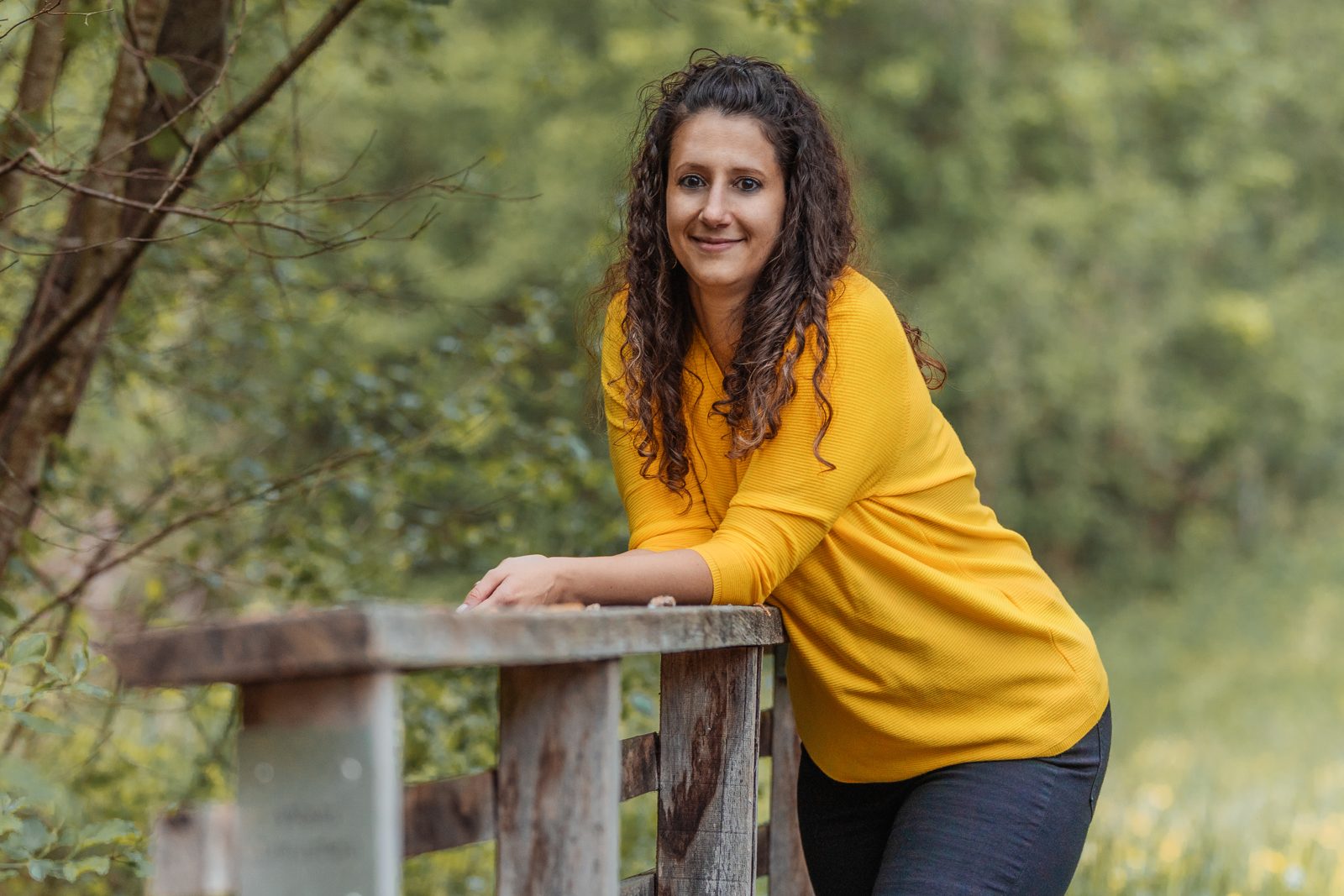 Sarah lehnt lächelnd an einem Holzgeländer im Grünen