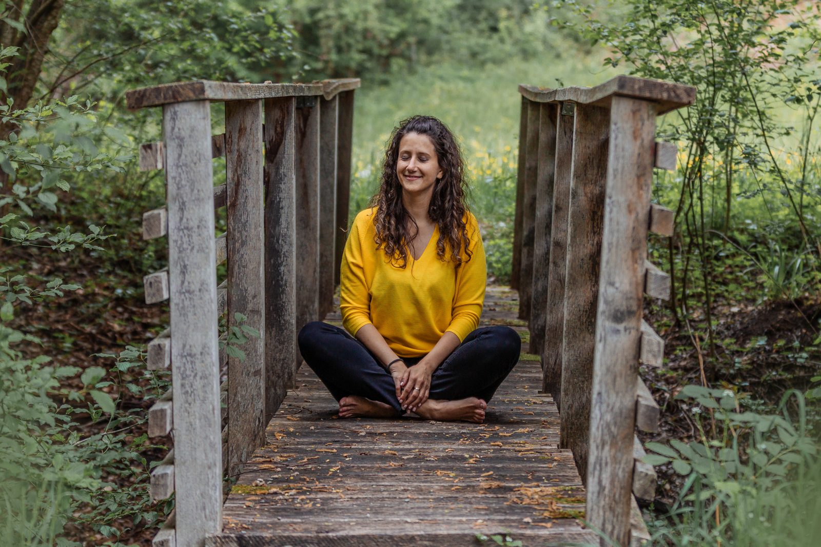 Sarah meditiert barfuss im Schneidersitz auf einer Holzbrücke im Wald